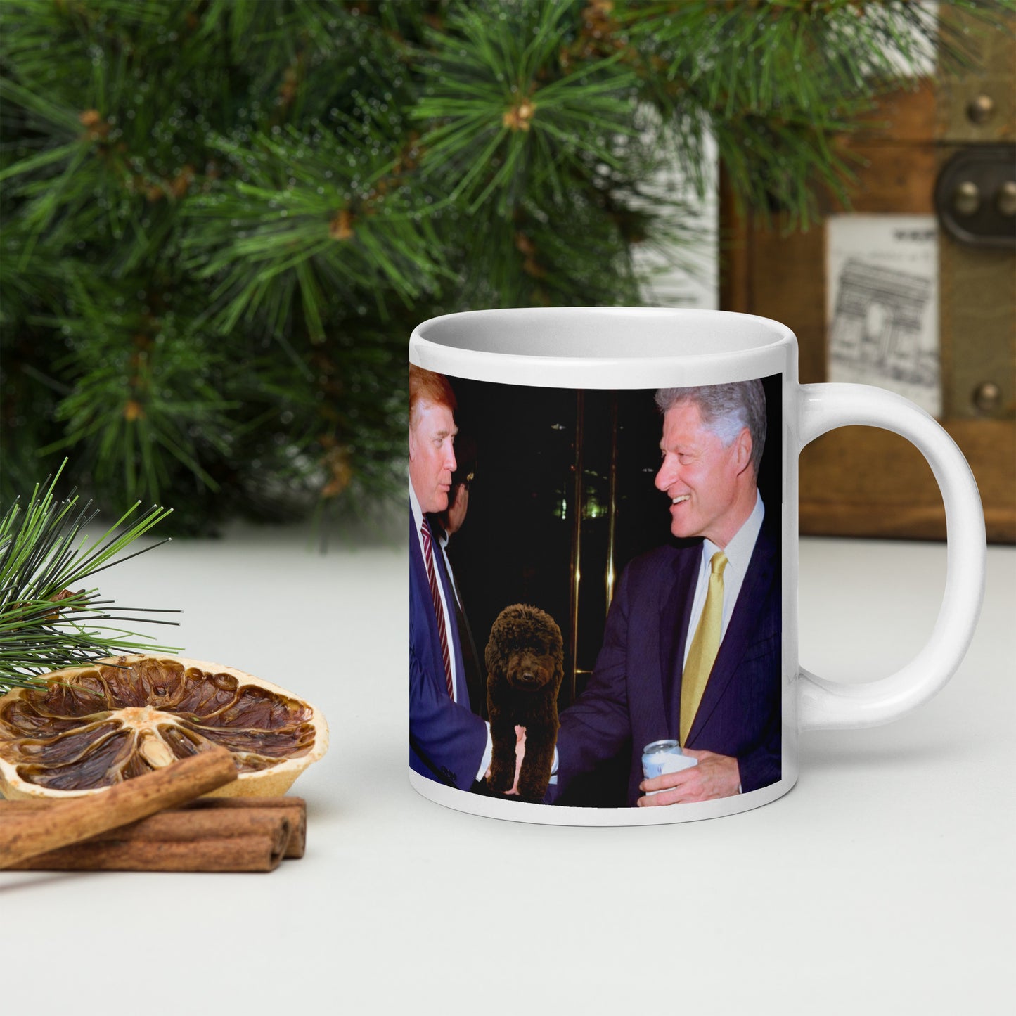 White glossy ceramic mug with a printed design of Bill Clinton and Donald Trump shaking hands at an event. A black dog is between them. Mug is on a white table with Christmas decorations around.