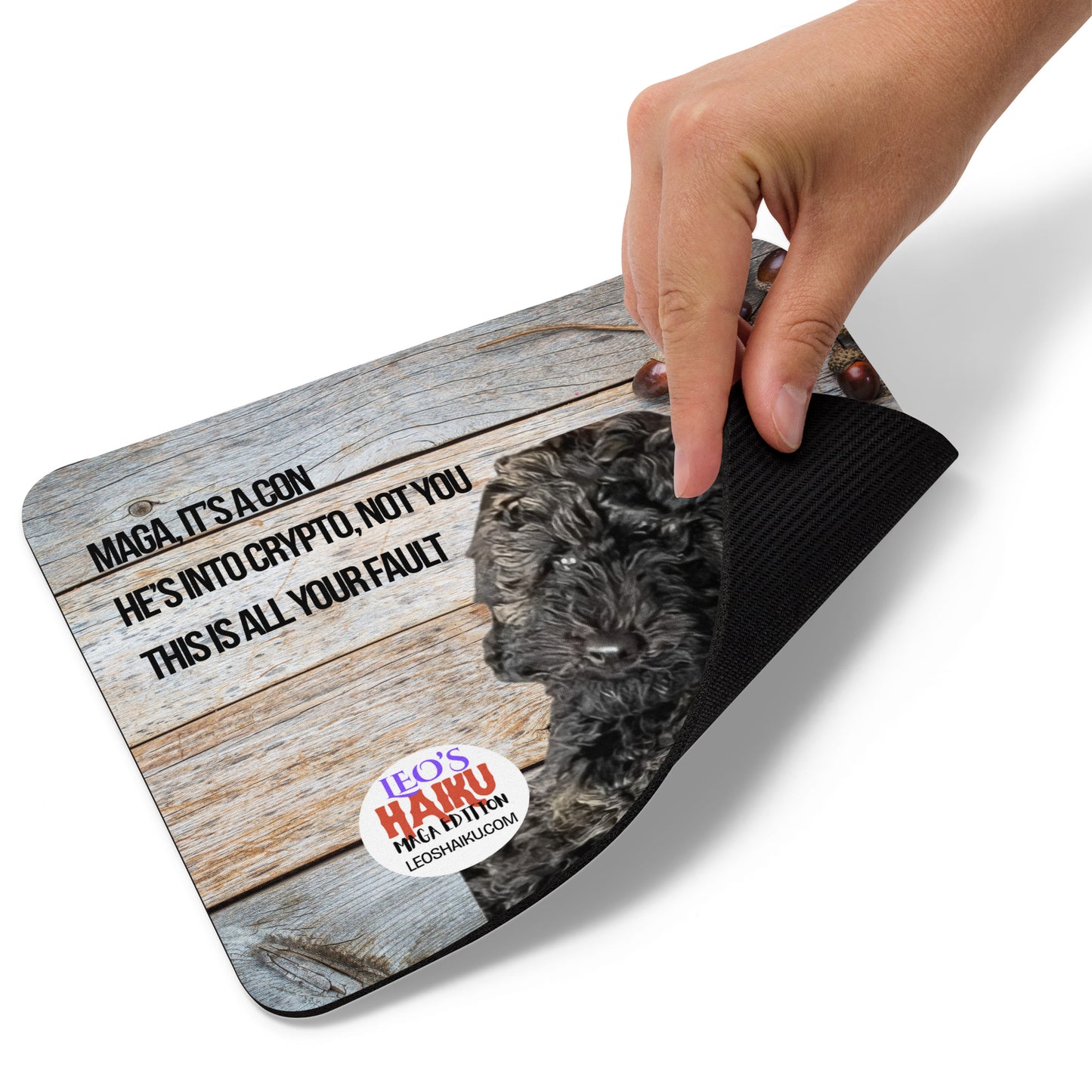 A hand lifts the bottom right edge of a standard mouse pad printed with the image of a dog against a natural wood background with acorns upper right. A political, satirical, anti-MAGA haiku reads: MAGA, It's a Con
He's into crypto, not you
This is all your fault
The text logo for leoshaiku.com is to the left of the dog. This view shows the thickness of the pad.