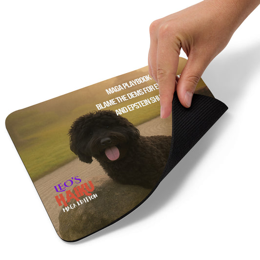 A hand lifts the right bottom edge of a mouse pad printed with an image of a black dog leaning against a rock in a zen garden. Political, satirical, anti-maga haiku is shown as is the Leoshaiku.com text logo. This view shows the thickness of the pad.