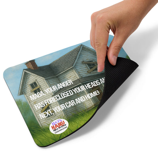 A hand lifts the lower right edge of a Mouse pad printed with the image of a boarded up house on an overgrown lawn. A small black dog sits at the front. Political, satirical, anti-Maga haiku is shown as is the text logo for leoshaiku.com. This view shows the thickness of the pad.