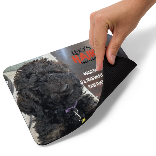 Hand lifts the lower right edge of a standard-sized mouse pad printed with a dog against a dark background and political satire in Haiku format. This view shows the thickness of the pad