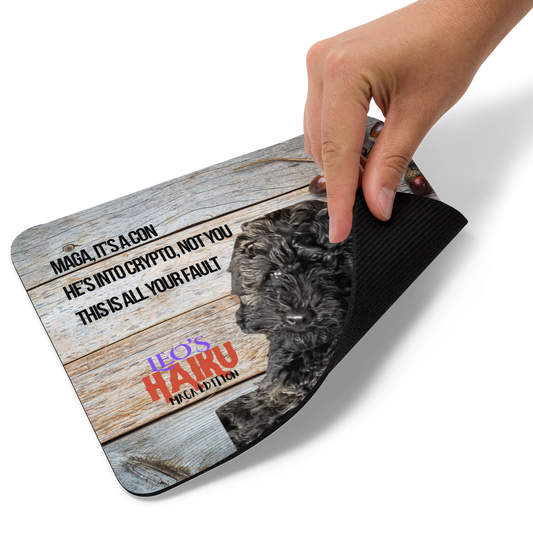 Hand lifting the corner of a standard-sized mouse pad printed with a dog against a wooden background and political satire in Haiku format. This view shows the thickness of the pad.