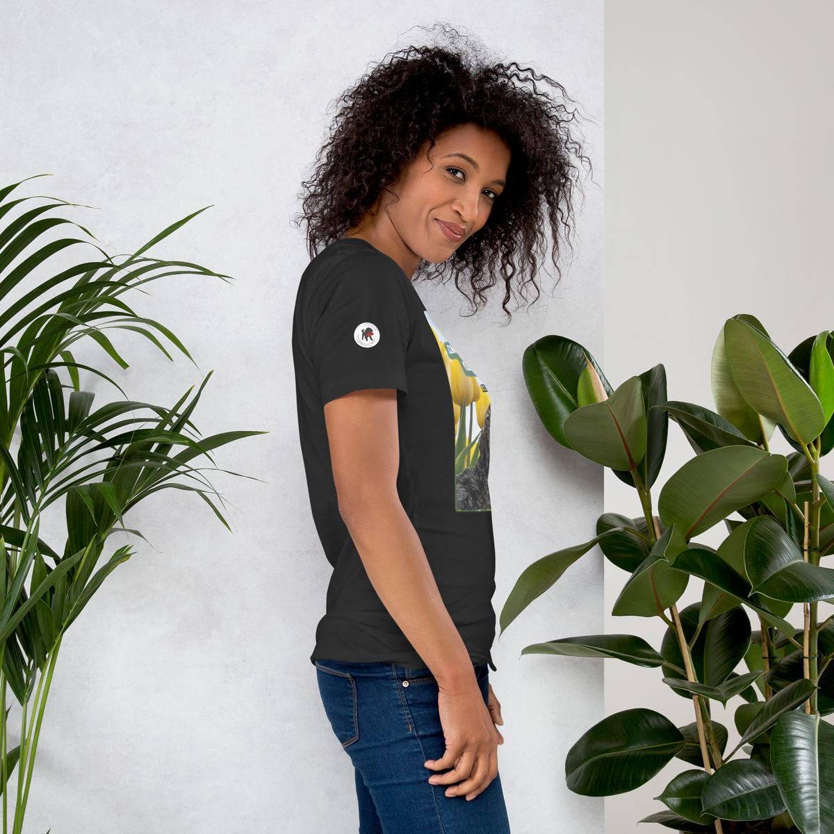 Woman wearing a dark grey t-shirt with a logo on the right sleeve, standing between two potted plants against a white wall.
