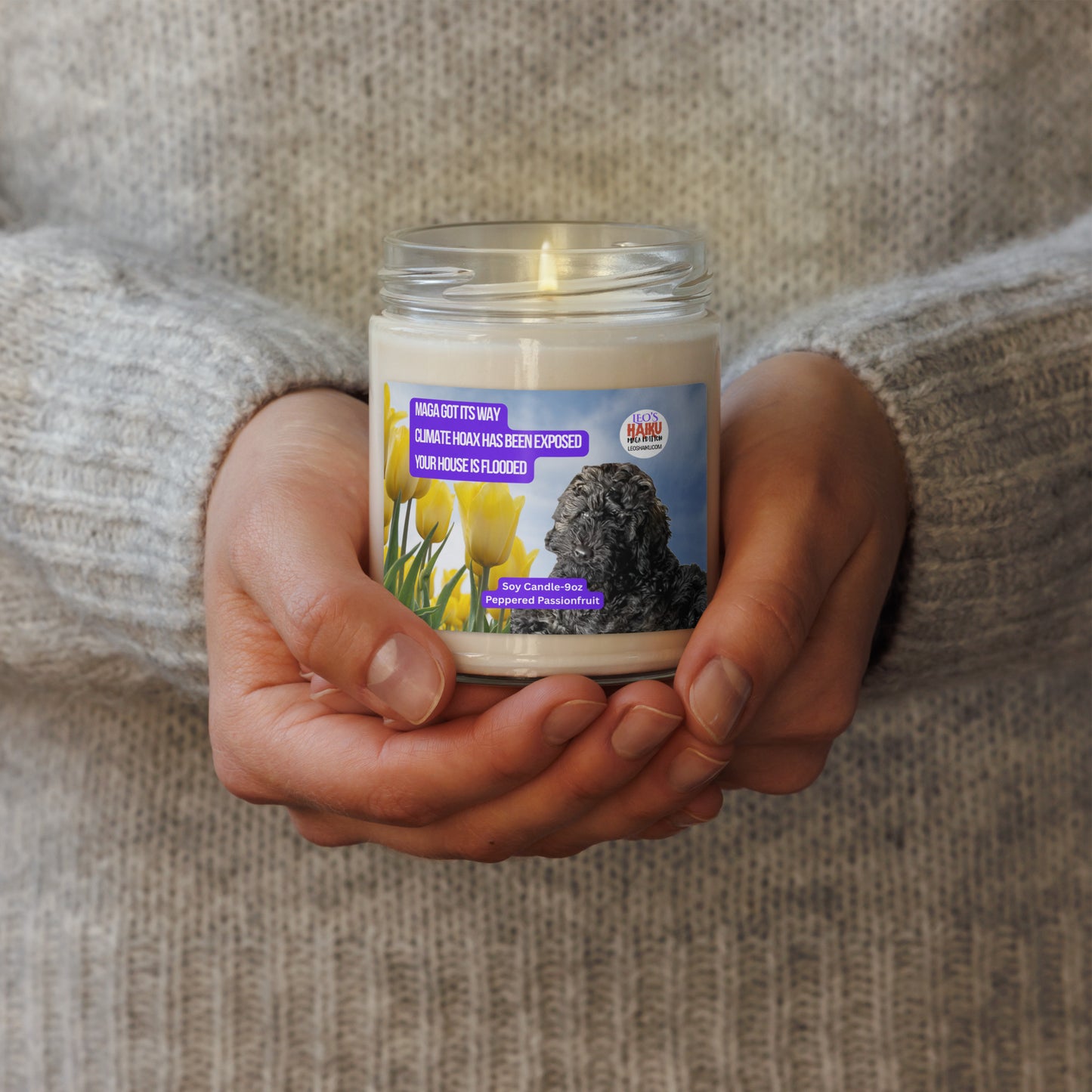 A person in a wheat-colored wool sweater holds a lighted candle in a glass jar.  The candle has a label that shows the image of a black dog lying near a group of yellow tulips. Top left is a political, satirical, anti-MAGA haiku, white text on a purple background, reads:  
MAGA got its way
Climate Hoax has been exposed
Your house is flooded.
Top right is the text logo for leoshaiku.com; Bottom center is another text bubble that reads Soy Candle 9 oz; Peppered Passionfruit