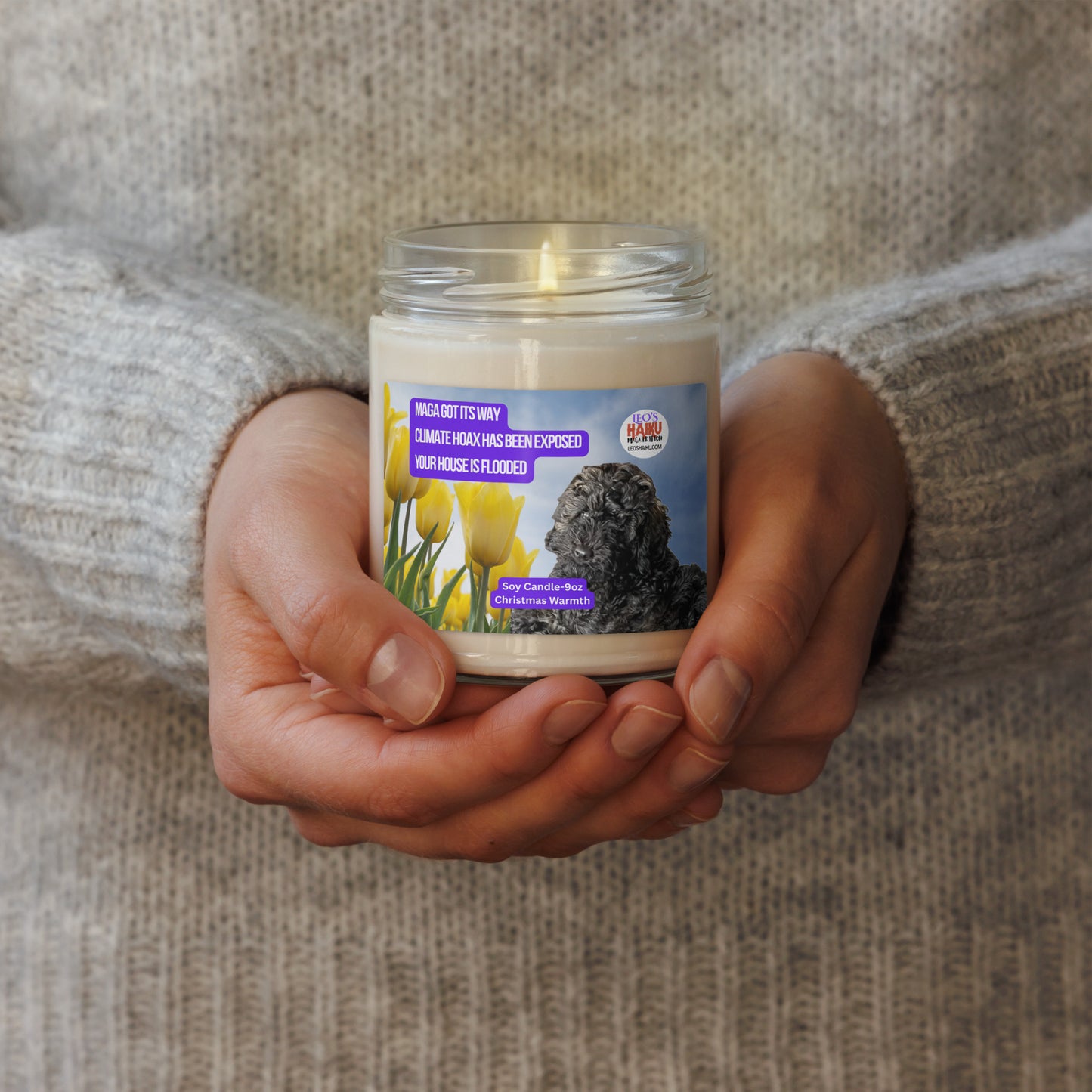 A person in a wheat-colored wool sweater holds a lighted candle in a glass jar.  The candle has a label that shows the image of a black dog lying near a group of yellow tulips. Top left is a political, satirical, anti-MAGA haiku, white text on a purple background, reads:  
MAGA got its way
Climate Hoax has been exposed
Your house is flooded.
Top right is the text logo for leoshaiku.com; Bottom center is another text bubble that reads Soy Candle 9 oz; Christmas Warmth