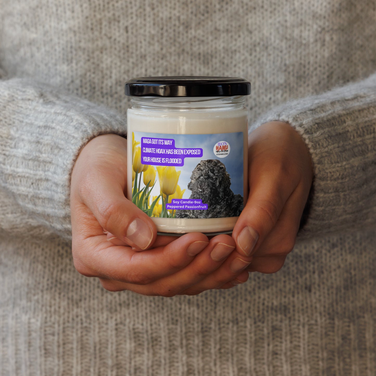 A person in a wheat-colored wool sweater holds a candle in a glass jar.  The candle has a label that shows the image of a black dog lying near a group of yellow tulips. Top left is a political, satirical, anti-MAGA haiku, white text on a purple background, reads:  
MAGA got its way
Climate Hoax has been exposed
Your house is flooded.
Top right is the text logo for leoshaiku.com; Bottom center is another text bubble that reads Soy Candle 9 oz; Peppered Passionfruit