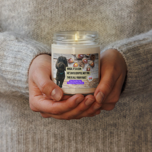 Person in a beige sweater holds a lighted candle in a glass jar.  The label shows a black dog superimposed on a horizontal wood plank background with acorns scattered top right.  In the center, the political, satirical, anti-MAGA haiku in black text with tan background is shown.  It reads:
MAGA It's a Con
He's Into Crypto, Not You
This is All Your Fault
Bottom center is white text on a purple background that reads Soy Candle - 9oz Peppered Passionfruit.
The text logo for leoshaiku.com is bottom right.