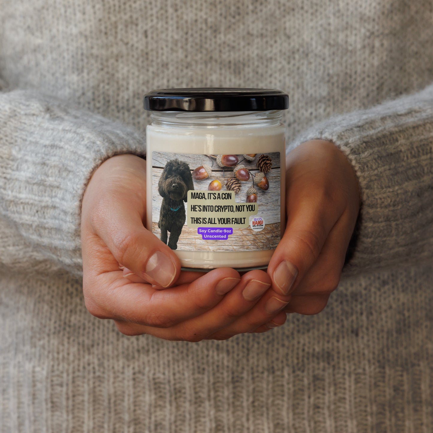 Person in a beige sweater holds a candle in a glass jar w/ lid.  The label shows a black dog superimposed on a horizontal wood plank background with acorns scattered top right.  In the center, the political, satirical, anti-MAGA haiku in black text with tan background is shown.  It reads:
MAGA It's a Con
He's Into Crypto, Not You
This is All Your Fault
Bottom center is white text on a purple background that reads Soy Candle - 9oz Unscented.
The text logo for leoshaiku.com is bottom right.