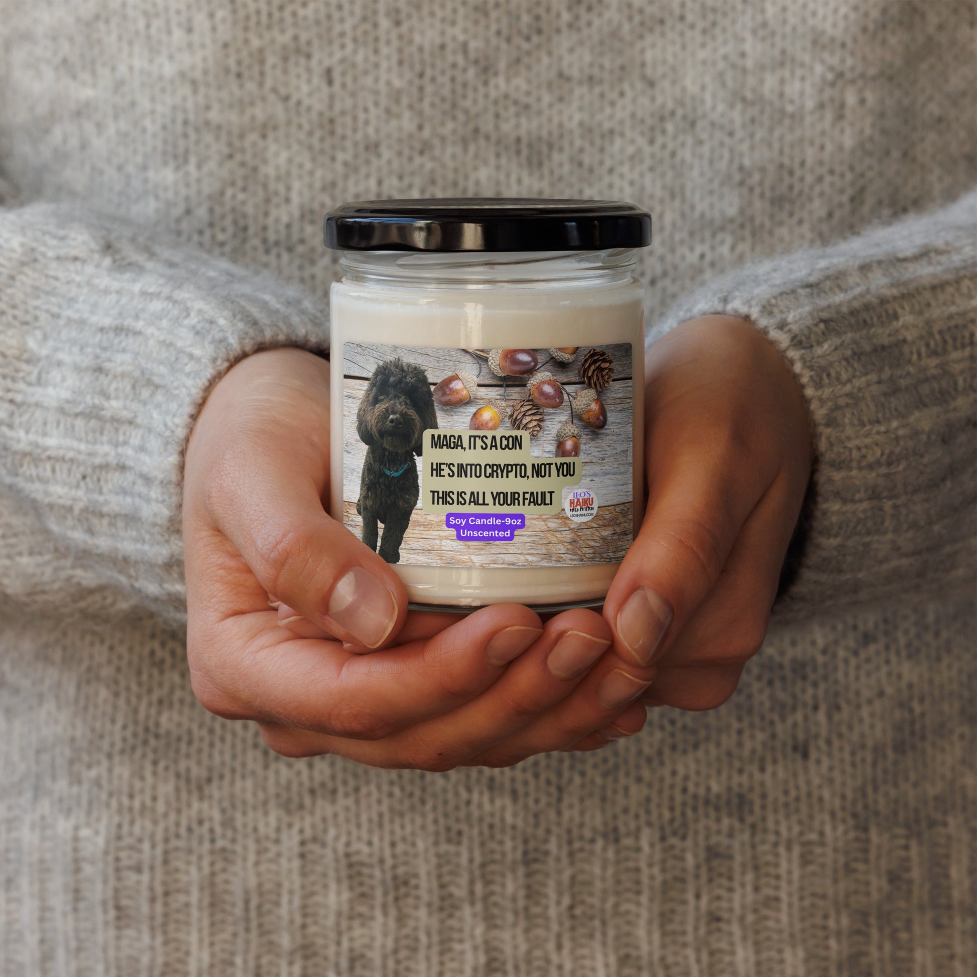 Person in a beige sweater holds a candle in a glass jar w/ lid.  The label shows a black dog superimposed on a horizontal wood plank background with acorns scattered top right.  In the center, the political, satirical, anti-MAGA haiku in black text with tan background is shown.  It reads:
MAGA It's a Con
He's Into Crypto, Not You
This is All Your Fault
Bottom center is white text on a purple background that reads Soy Candle - 9oz Unscented.
The text logo for leoshaiku.com is bottom right.