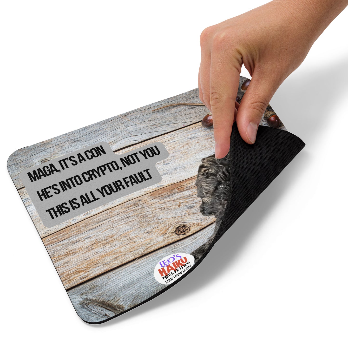 A hand lifts the bottom right corner of a standard Mouse Pad with an image of Leo the Black Mini Poodle top left against a wooden background.  Acorns and pine cones top right with the round text logo for leoshaiku.com below.  Centered is a political, satirical, anti-MAGA haiku that reads:  MAGA, It's a Con
He's Into Crypto, Not You
This is All Your Fault

This view shows the thickness of the pad.