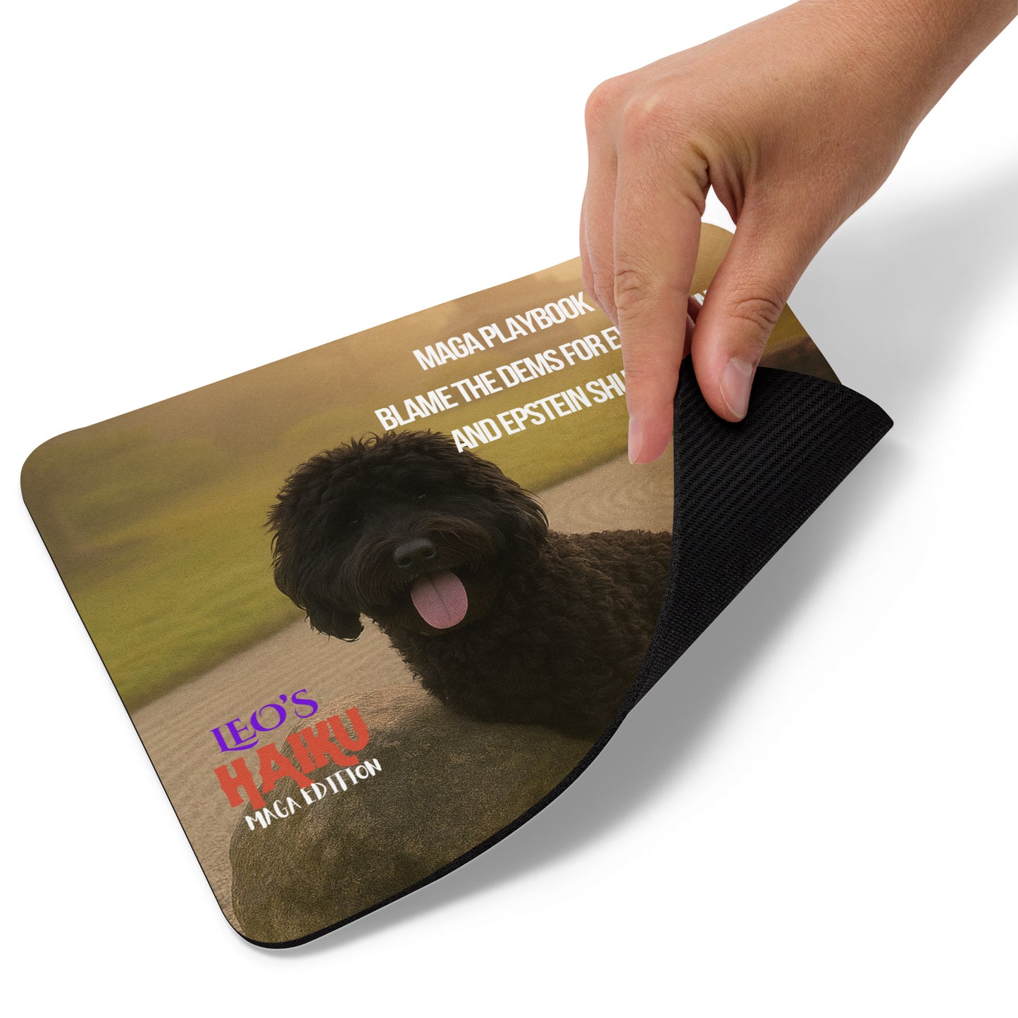 A hand lifts the right bottom edge of a mouse pad  printed with an image of a black dog leaning against a rock in a zen garden.  Political, satirical, anti-maga haiku is shown as is the Leoshaiku.com text logo.  This view shows the thickness of the pad.