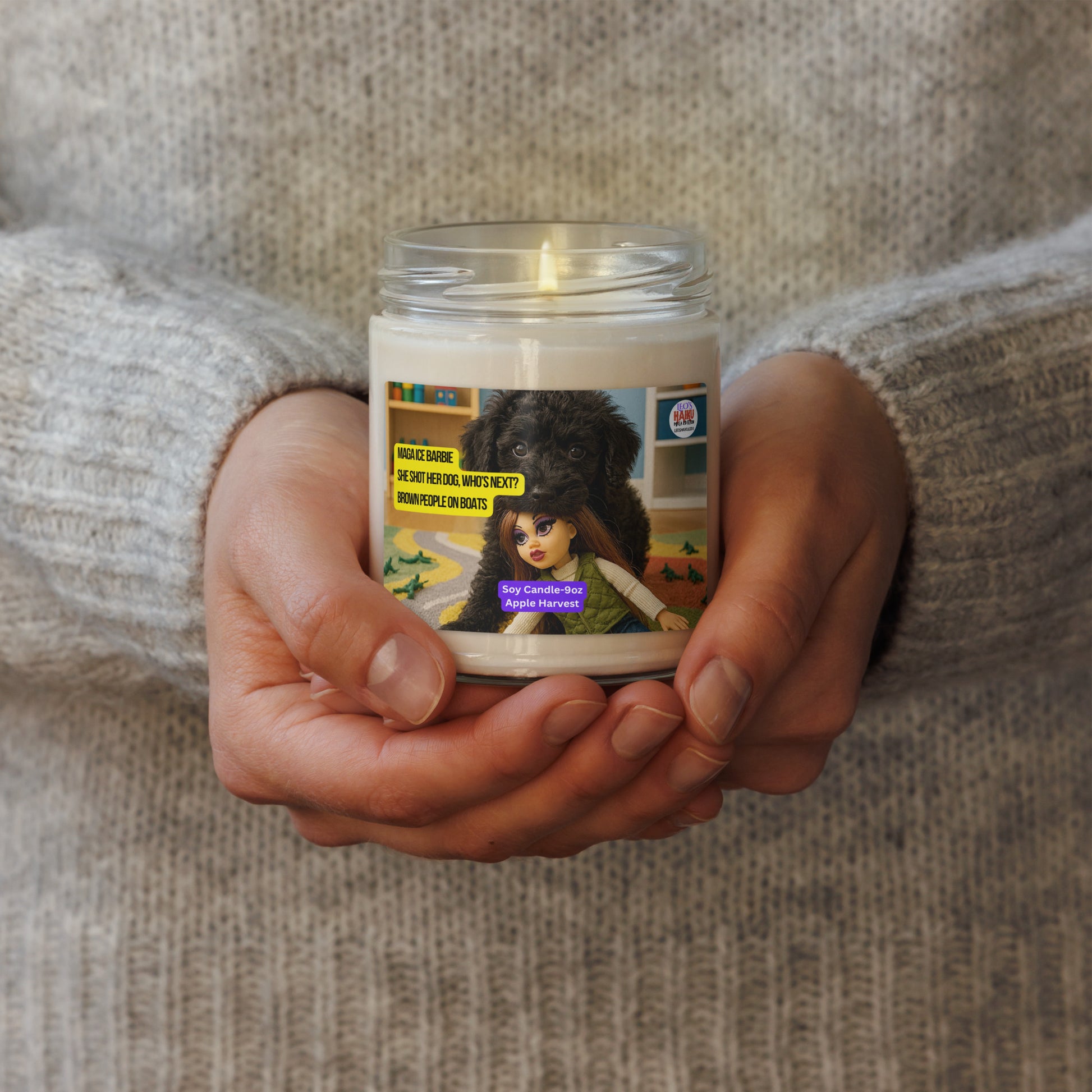 2 hands hold a lighted candle in a glass jar.  The label shows a black dog in a child's playroom.  Dog has a fashion dolls head in his mouth.  Text logo for leoshaiku.com is upper right.  A political, satirical, anti-MAGA haiku is displayed that reads:  MAGA ICE Barbie; She Shot Her Dog, Who's Next?, Brown People on Boats.  Bottom Center is text that reads Soy Candle - 9 oz, Apple Harvest