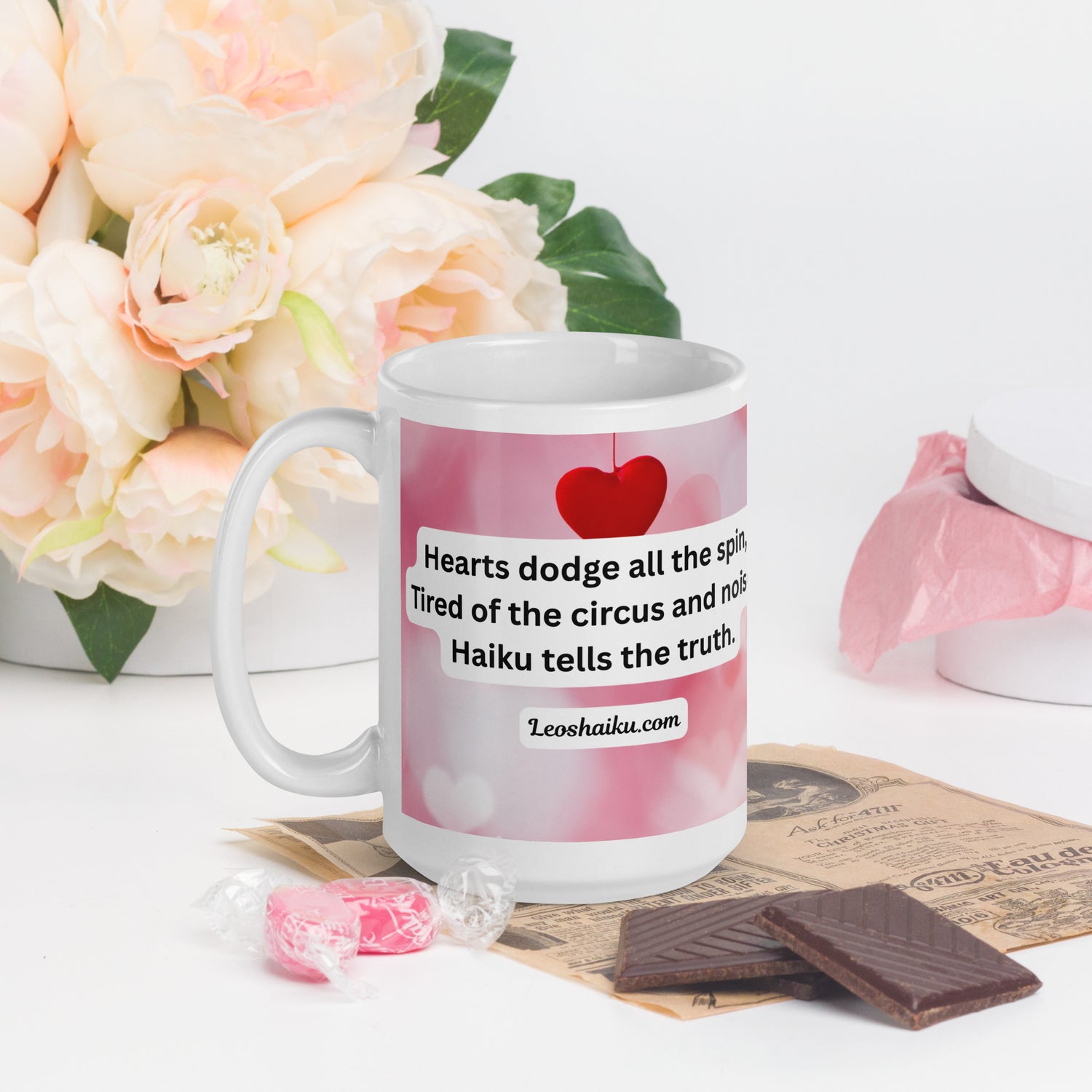 A white ceramic mug printed with a background of pink and valentine hearts is displayed in a Valentine-themed backdrop of flowers and candy.  Handle on the left.  The haiku reads 
Hearts dodge all the spin,
Tired of the circus and noise,
Haiku tells the truth.
Underneath it says leoshaiku.com
