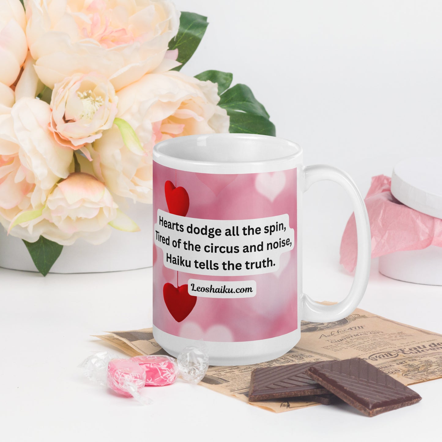 A white ceramic mug printed with a background of pink and valentine hearts is displayed in a Valentine-themed backdrop of flowers and candy.  Handle on the right. The haiku reads 
Hearts dodge all the spin,
Tired of the circus and noise,
Haiku tells the truth.
Underneath it says leoshaiku.com