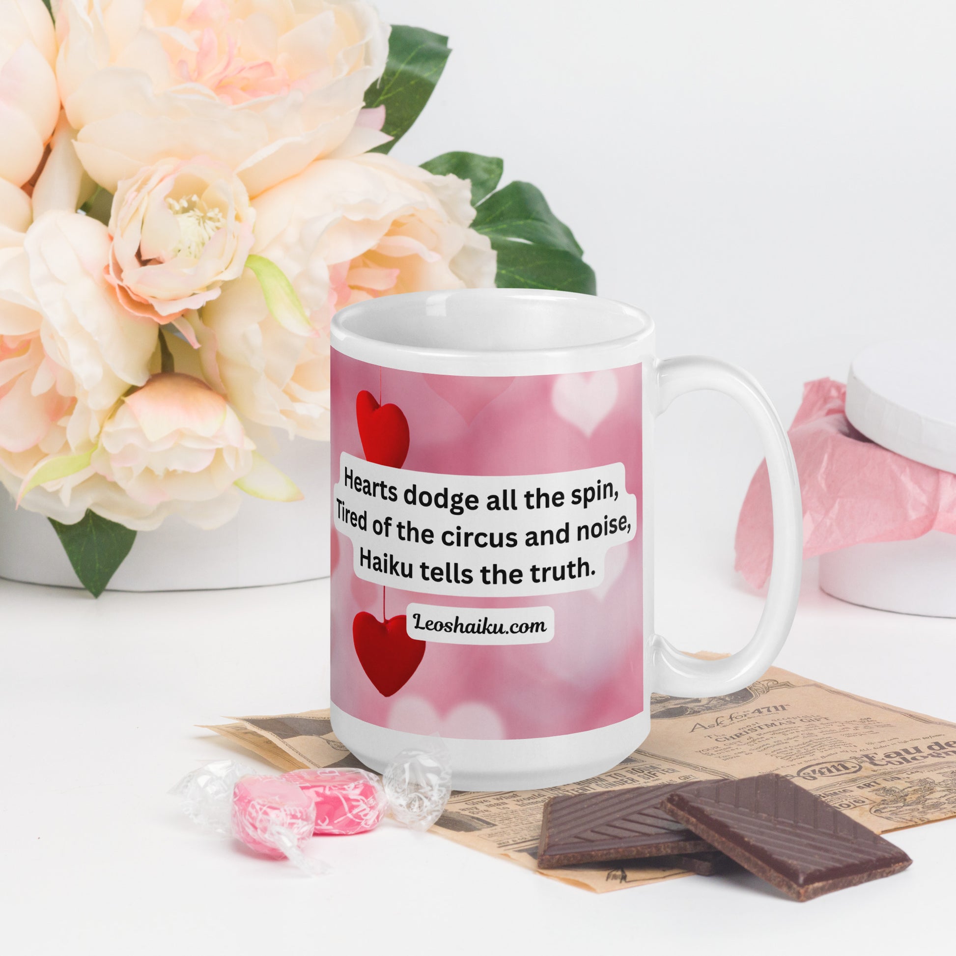 A white ceramic mug printed with a background of pink and valentine hearts is displayed in a Valentine-themed backdrop of flowers and candy.  Handle on the right. The haiku reads 
Hearts dodge all the spin,
Tired of the circus and noise,
Haiku tells the truth.
Underneath it says leoshaiku.com