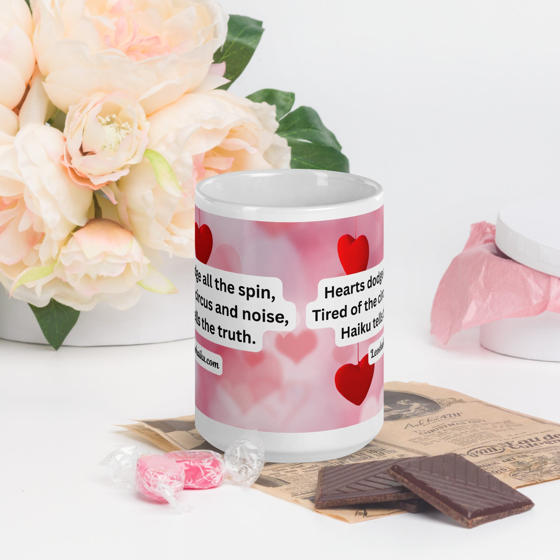 A white ceramic mug printed with a background of pink and valentine hearts is displayed in a Valentine-themed backdrop of flowers and candy.  The haiku reads 
Hearts dodge all the spin,
Tired of the circus and noise,
Haiku tells the truth.
Underneath it says leoshaiku.com