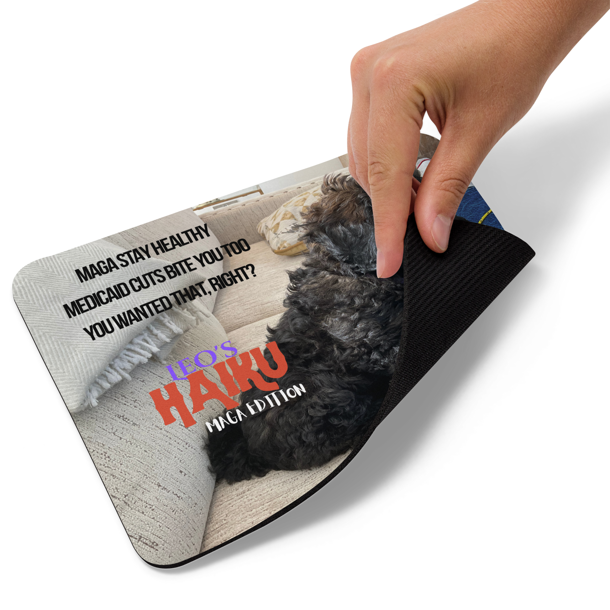 A hand lists the lower right corner of a standard-sized mouse pad printed with the side view of a poodle sitting on a beige couch, political satire in Haiku format, and branding elements.  This view shows the thickness of the pad.  