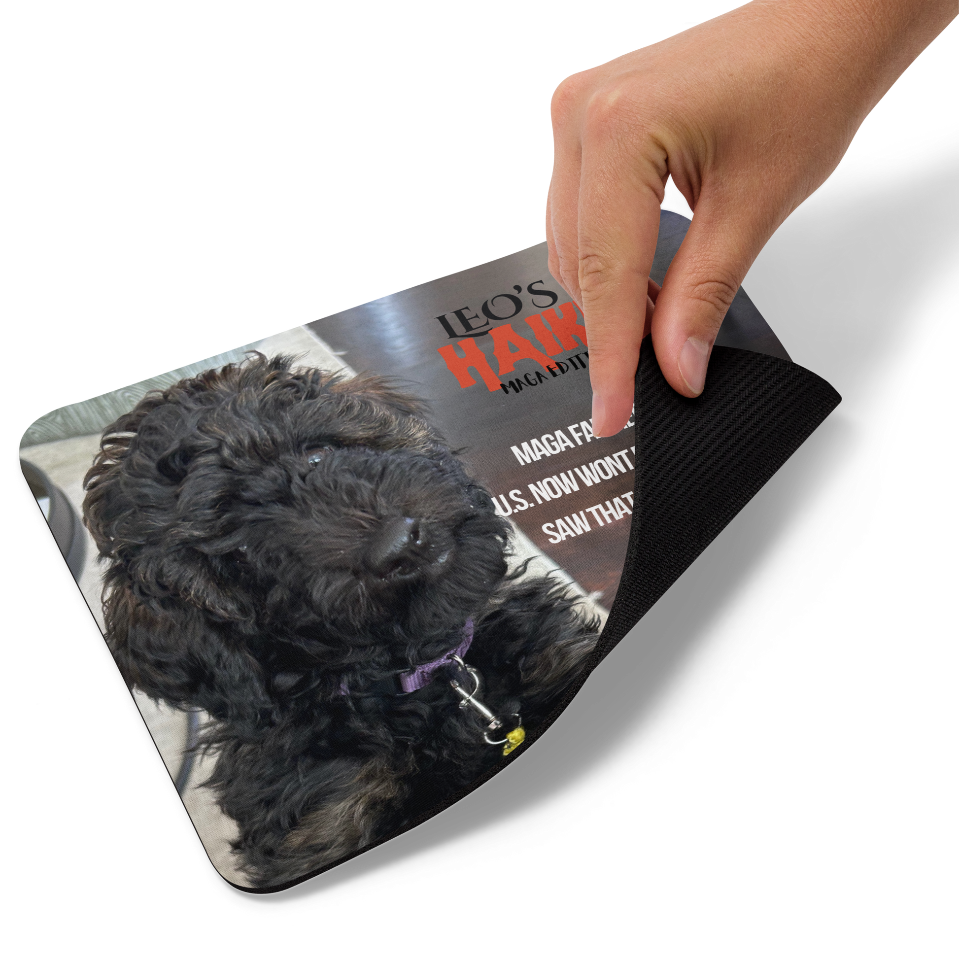 Hand lifts the lower right edge of a standard-sized mouse pad printed with a dog against a dark background and political satire in Haiku format.  This view shows the thickness of the pad