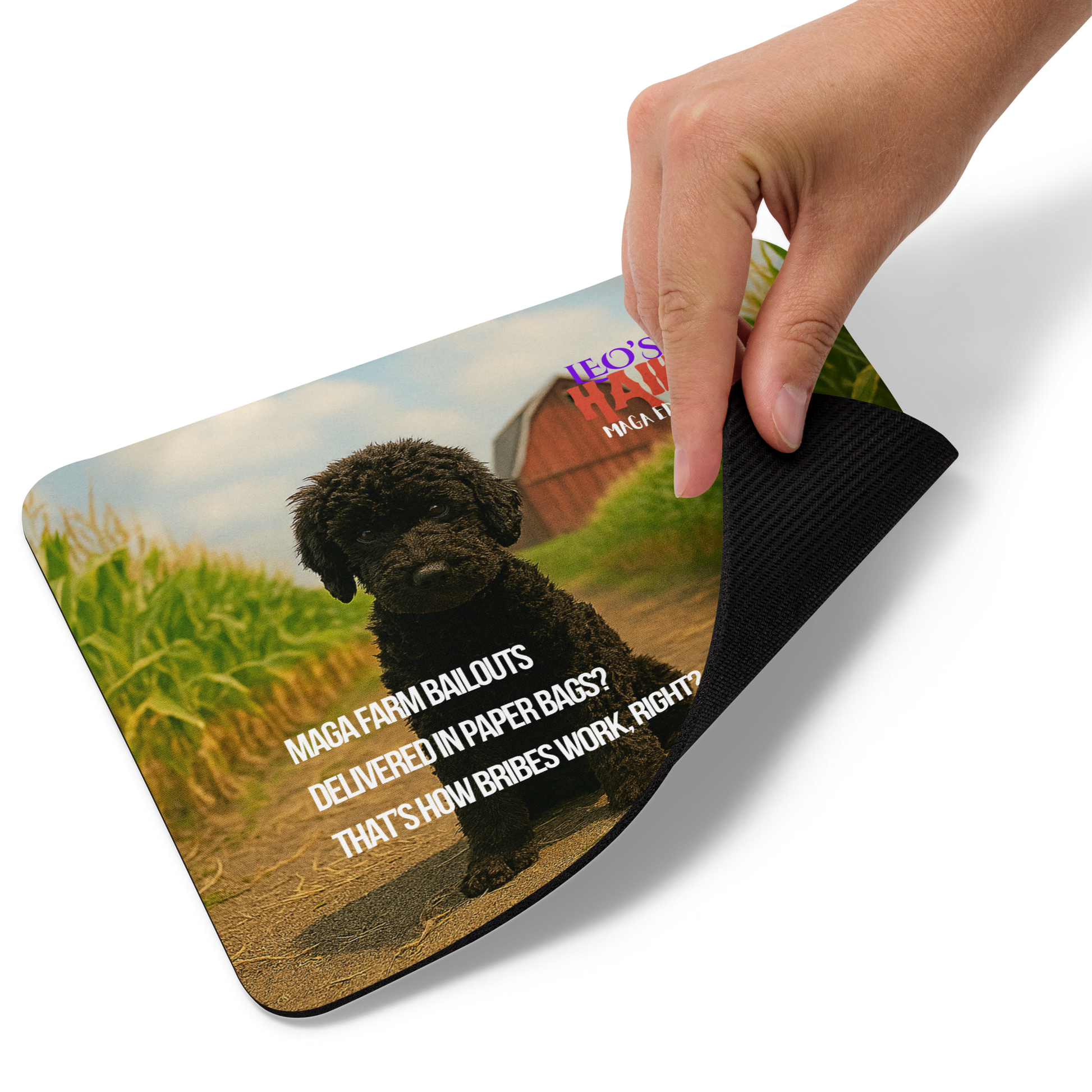 Hand is lifting the lower right corner of a standard-sized mouse pad printed with a barn/cornfield scene, a dog and political satire in Haiku format.  This view shows the thickness of the pad