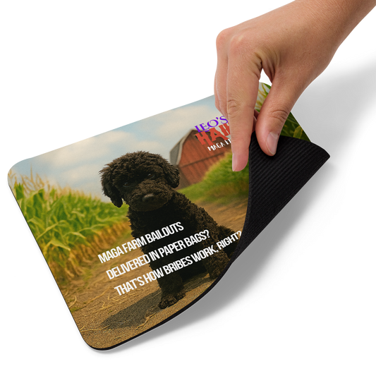 Hand is lifting the lower right corner of a standard-sized mouse pad printed with a barn/cornfield scene, a dog and political satire in Haiku format.  This view shows the thickness of the pad