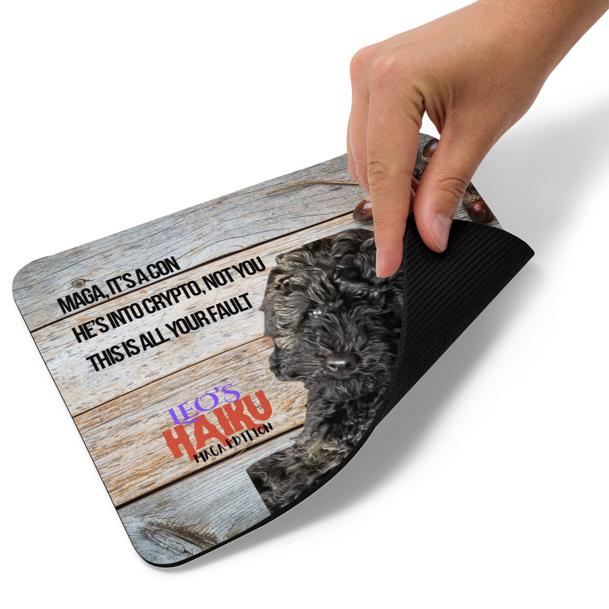 Hand lifting the corner of a standard-sized mouse pad printed with a dog against a wooden background and political satire in Haiku format. This view shows the thickness of the pad.