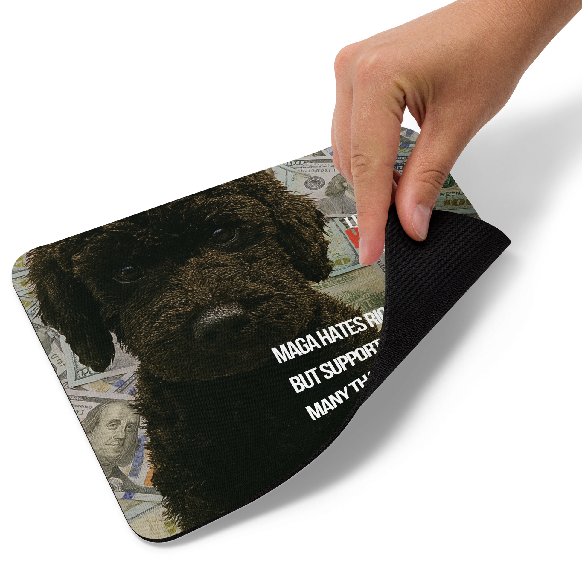 A hand lifts the bottom right corner of a standard-sized mouse pad printed with the large face of black dog with $100 bills behind him; political satire text and brand.  This view shows the thickness of the pad.