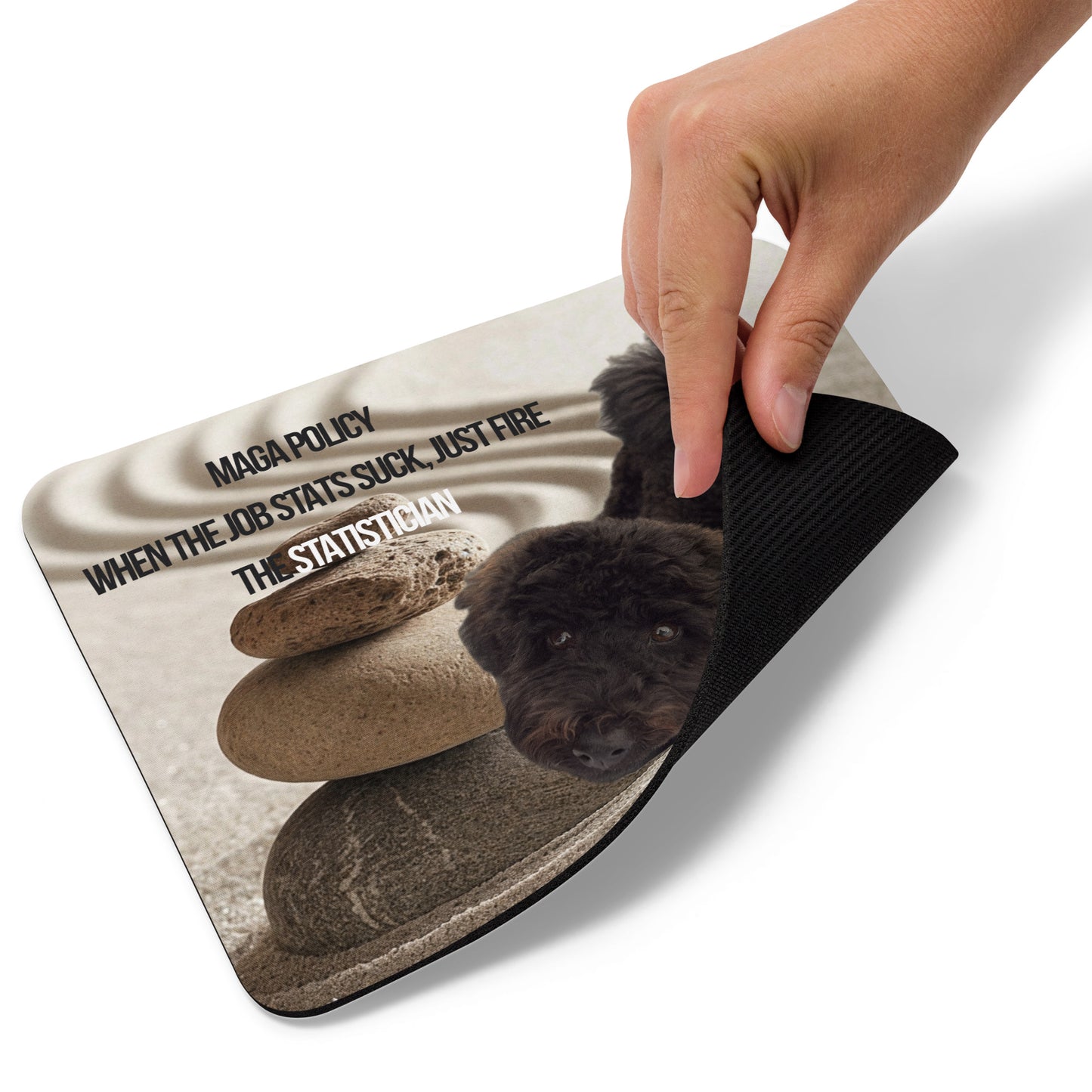 A hand lifts the lower right edge of a standard-sized mouse pad printed with the image of a black dog standing in a Zen garden with sand and a stack of rocks, political satire text and branding.  This view shows the thickness of the pad.