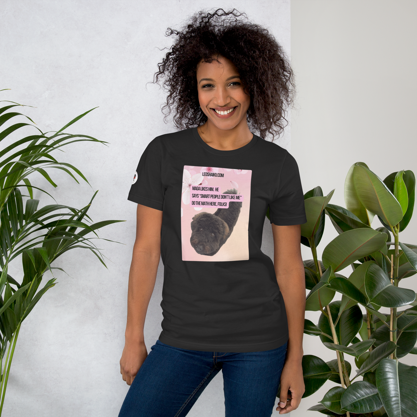 Woman facing front in a dark gray t-shirt.  It's printed with the image of a black dog looking into the camera from a pink flowered background.  A political, satirical, anti-maga haiku reads:
MAGA likes him.  He
says “smart people don’t like me”
do the math here, FOLKS!