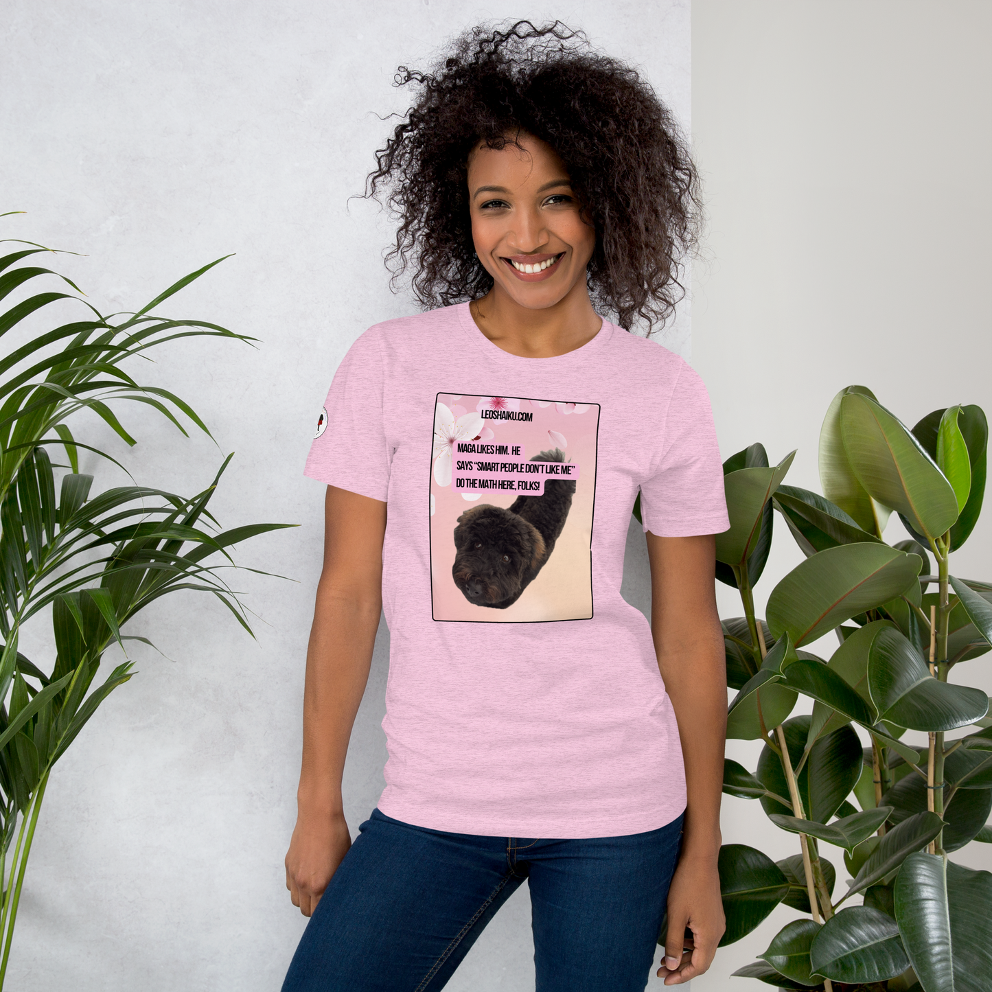 Woman facing front in a heather prism lilac t-shirt.  It's printed with the image of a black dog looking into the camera from a pink flowered background.  A political, satirical, anti-maga haiku reads:
MAGA likes him.  He
says “smart people don’t like me”
do the math here, FOLKS!