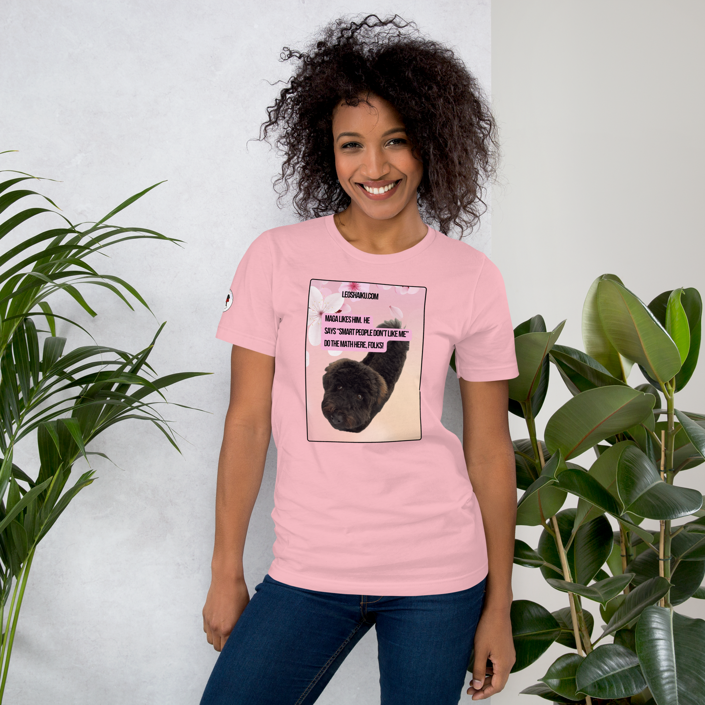Woman facing front in pink t-shirt.  It's printed with the image of a black dog looking into the camera from a pink flowered background.  A political, satirical, anti-maga haiku reads:
MAGA likes him.  He
says “smart people don’t like me”
do the math here, FOLKS!