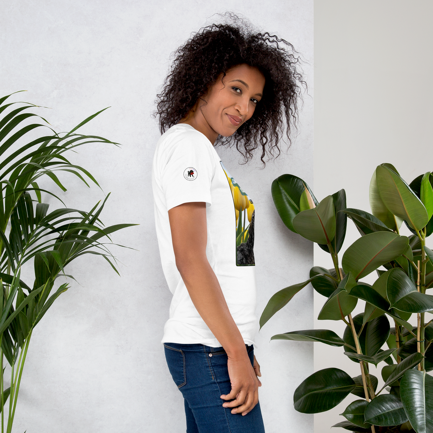 Woman wearing a white t-shirt with a logo on the right sleeve, standing between two potted plants against a white wall.